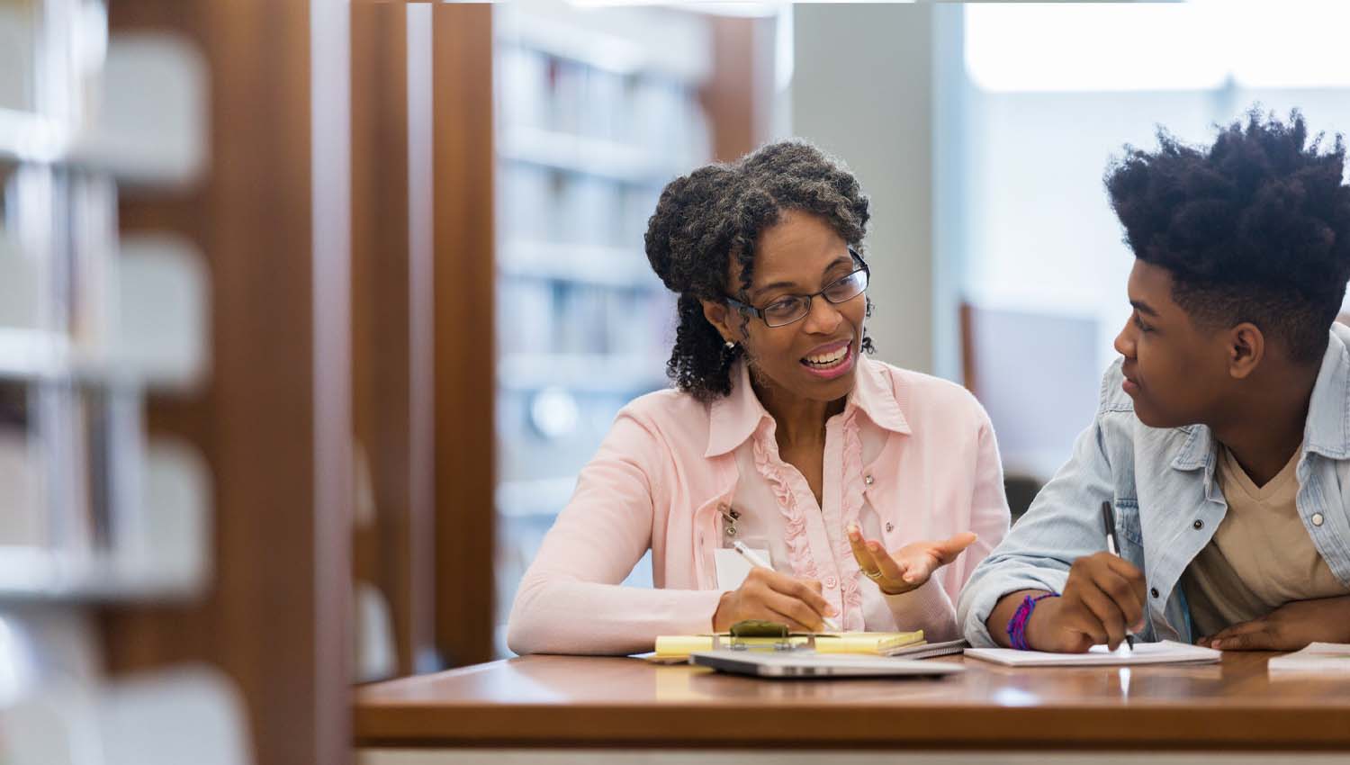 Teacher mentoring a student in a library setting, both engaged in discussion