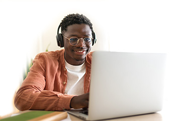 A college student sitting a desk with headphones on using a laptop and smiling