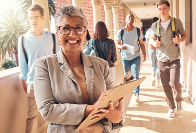 Happy school administrator holding a clipboard in a busy high school hallway.